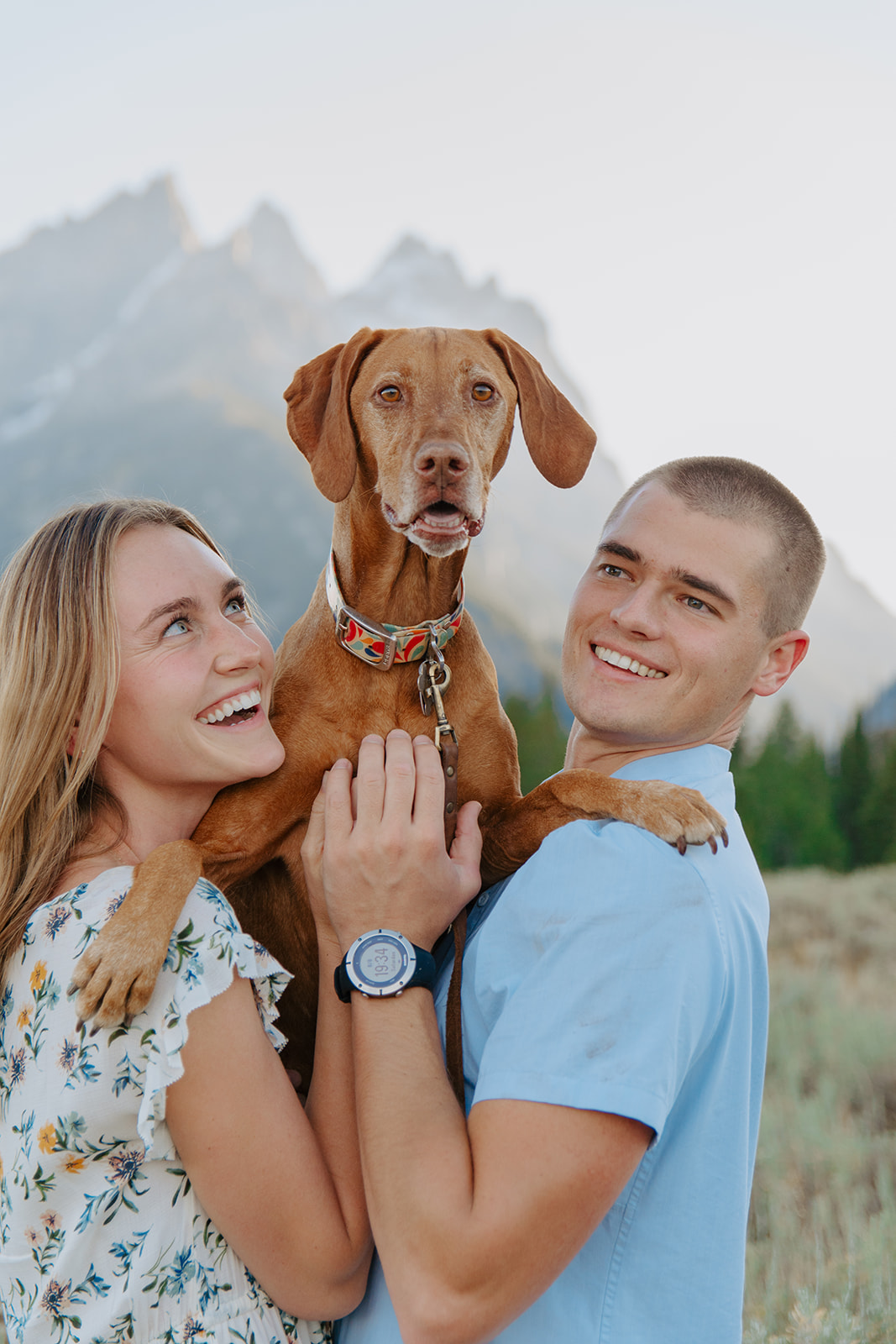 Couple stands in front of the Tetons with their dog.for engagement photos